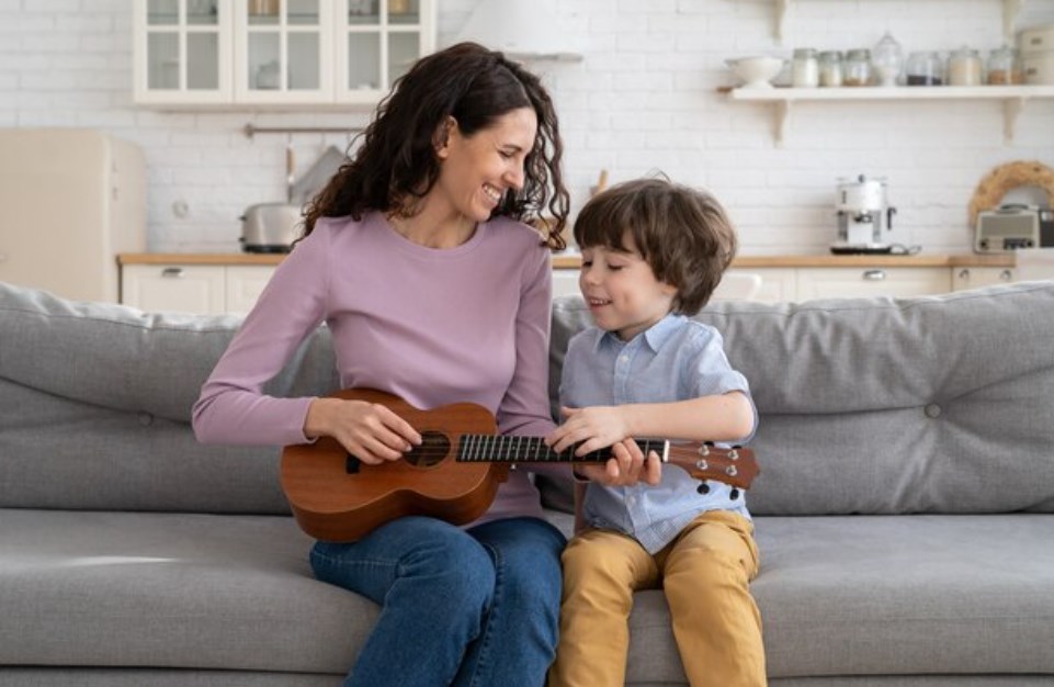 a young mother holding a ukulele while sitting on a couch with her little son